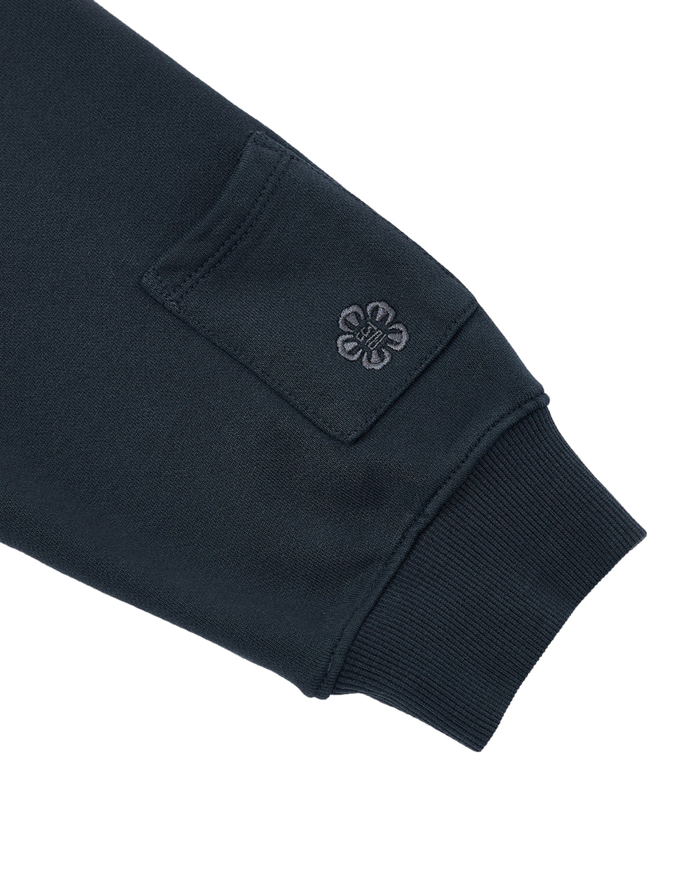 A close-up of the INDIGO WASH HANBOK HOODIE by Sundae School shows a dark sleeve with a ribbed cuff, highlighted by the signature sleeve stash pocket and an embroidered flower emblem just below it, taking inspiration from traditional Hanbok detailing.