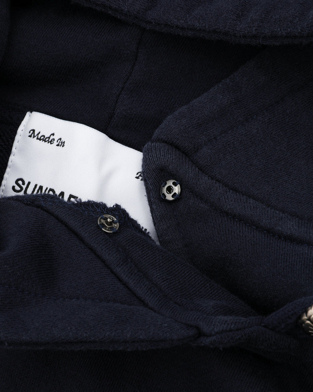 A close-up of the NAVY WASH HANBOK HOODIE by Sundae School shows its oversized hood, metal snap button, and a partially visible white label reading "Made in" and "SUNDA." Made from custom cotton, the fabric looks soft and thick.
