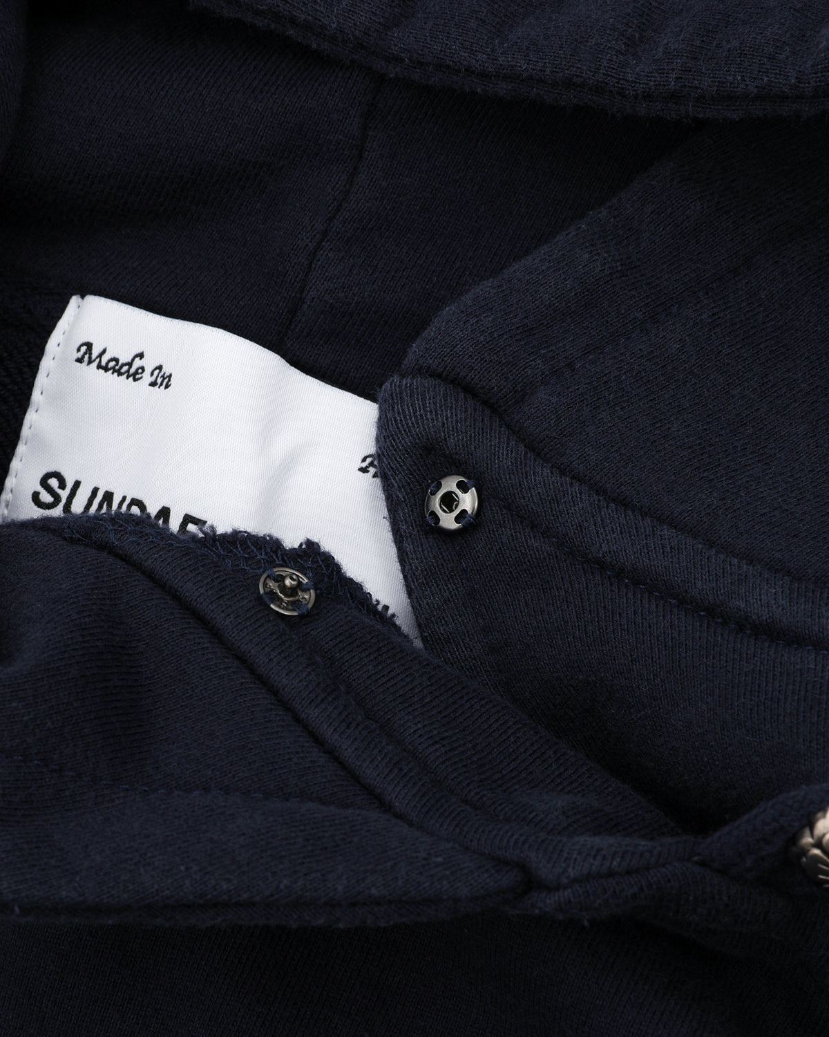 A close-up of the NAVY WASH HANBOK HOODIE by Sundae School shows its oversized hood, metal snap button, and a partially visible white label reading "Made in" and "SUNDA." Made from custom cotton, the fabric looks soft and thick.
