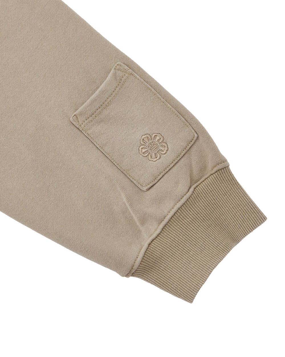A close-up of the TAUPE HANBOK HOODIE by Sundae School, showcasing its beige sleeve with a ribbed cuff and a small rectangular signature stash pocket adorned with an embroidered flower-like design.