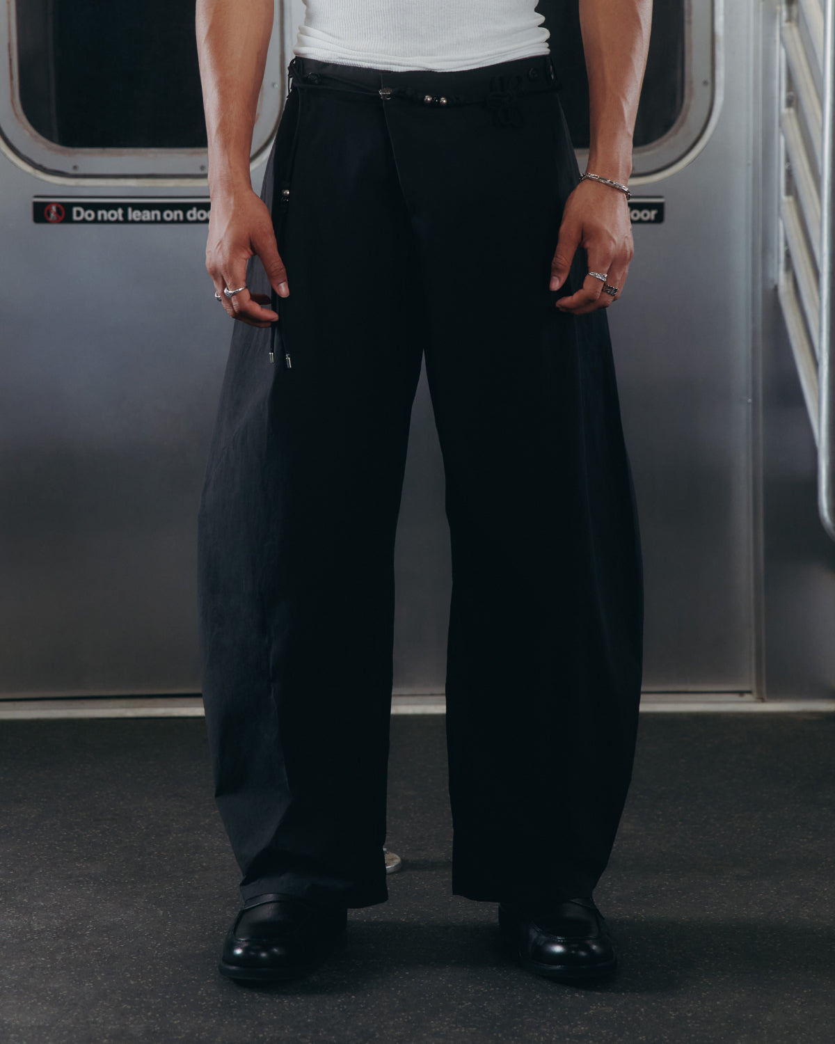 A person wearing Sundae School BLACK HANBOK LINEN PANTS, a white tank top, black shoes, and rings stands inside a subway car with their hands at their sides. The subway door and a safety sign appear in the background.
