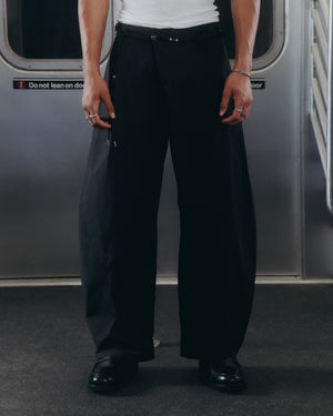 A person wearing Sundae School BLACK HANBOK LINEN PANTS, a white tank top, black shoes, and rings stands inside a subway car with their hands at their sides. The subway door and a safety sign appear in the background.