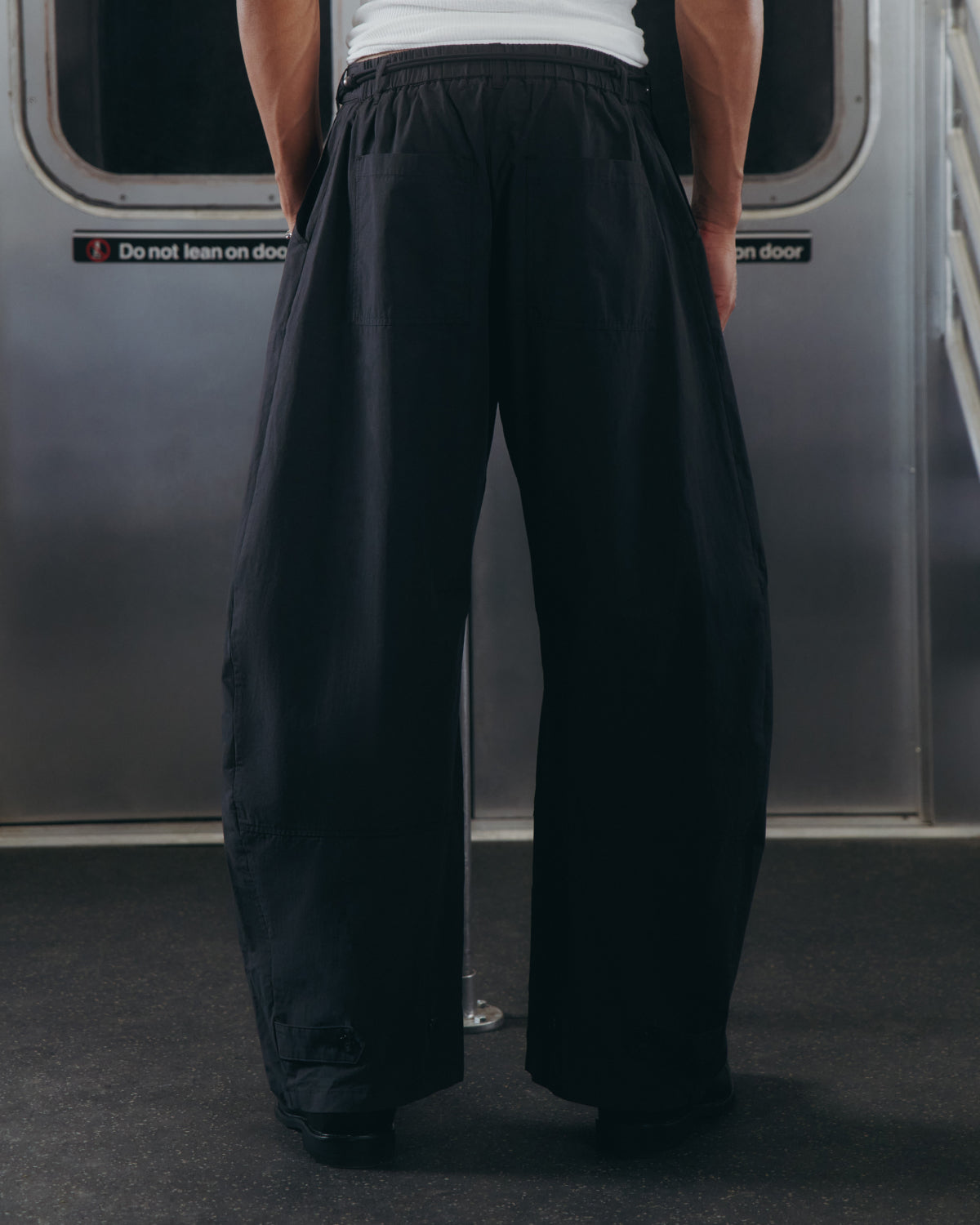 A person stands in a subway car, facing away from the camera, wearing Sundae School BLACK HANBOK LINEN PANTS and a white shirt. A sign on the door reads, Do not lean on door.