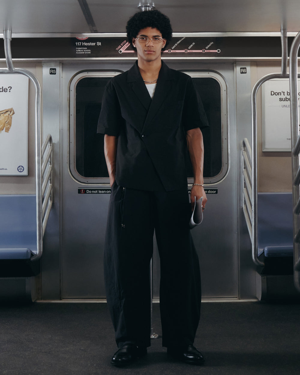 A person with curly hair and glasses stands in the middle of an empty subway car, wearing the BLACK HANBOK LINEN SHIRT by Sundae School. Subway seats and metal poles are visible, and they are holding a white item.