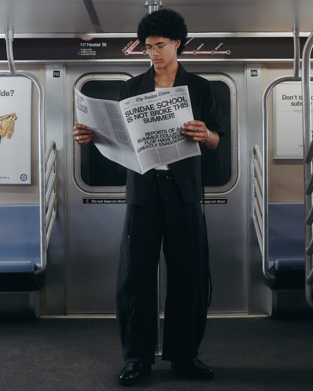 A person with curly hair and glasses, wearing Sundae School BLACK HANBOK LINEN PANTS, stands in a subway car reading a large newspaper headlined “SUNDAE SCHOOL IS NOT BROKE THIS SUMMER.” The subway stop displays 117 Hester St.