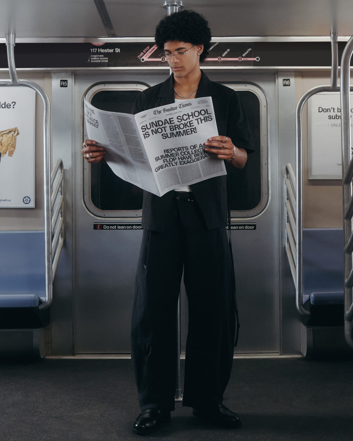 A person with curly hair and glasses, wearing Sundae School BLACK HANBOK LINEN PANTS, stands in a subway car reading a large newspaper headlined “SUNDAE SCHOOL IS NOT BROKE THIS SUMMER.” The subway stop displays 117 Hester St.