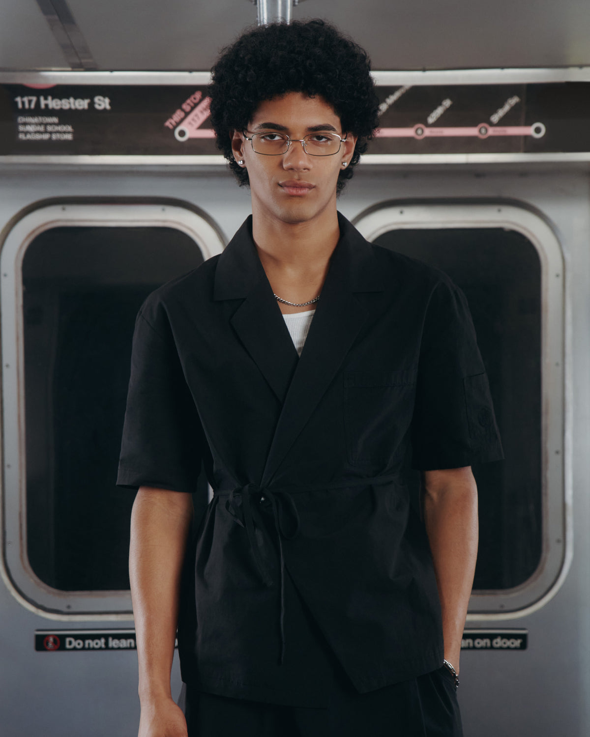 A young person with curly hair and glasses wears the BLACK HANBOK LINEN SHIRT by Sundae School in a subway car, standing beneath a 117 Hester St sign with a subway map beside it.