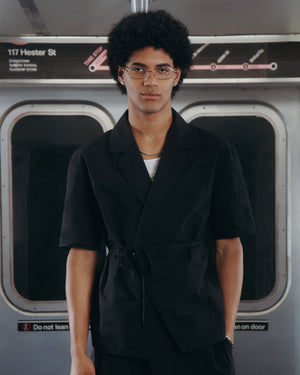 A young person with curly hair and glasses wears the BLACK HANBOK LINEN SHIRT by Sundae School in a subway car, standing beneath a 117 Hester St sign with a subway map beside it.