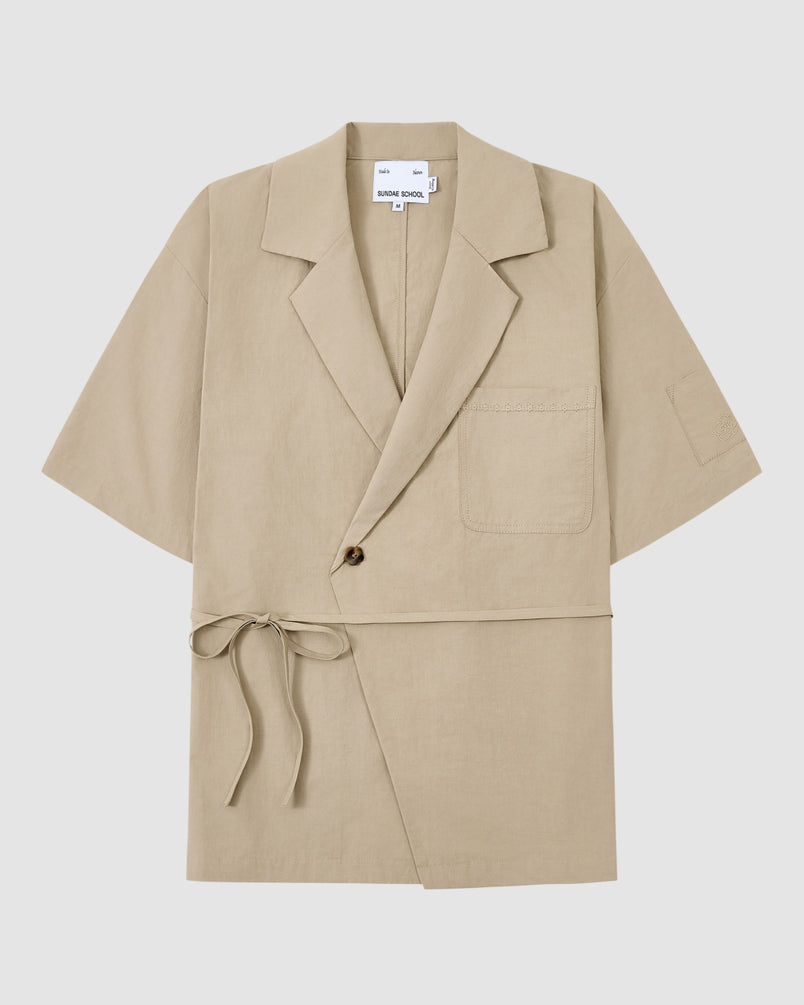 The WHEAT HANBOK LINEN SHIRT by Sundae School is a beige short-sleeve wrap jacket made from breathable cotton-linen blend, with a notched lapel, chest pocket, single-button closure, and side tie at the waist. Shown on a plain white background.