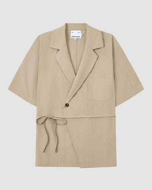 The WHEAT HANBOK LINEN SHIRT by Sundae School is a beige short-sleeve wrap jacket made from breathable cotton-linen blend, with a notched lapel, chest pocket, single-button closure, and side tie at the waist. Shown on a plain white background.