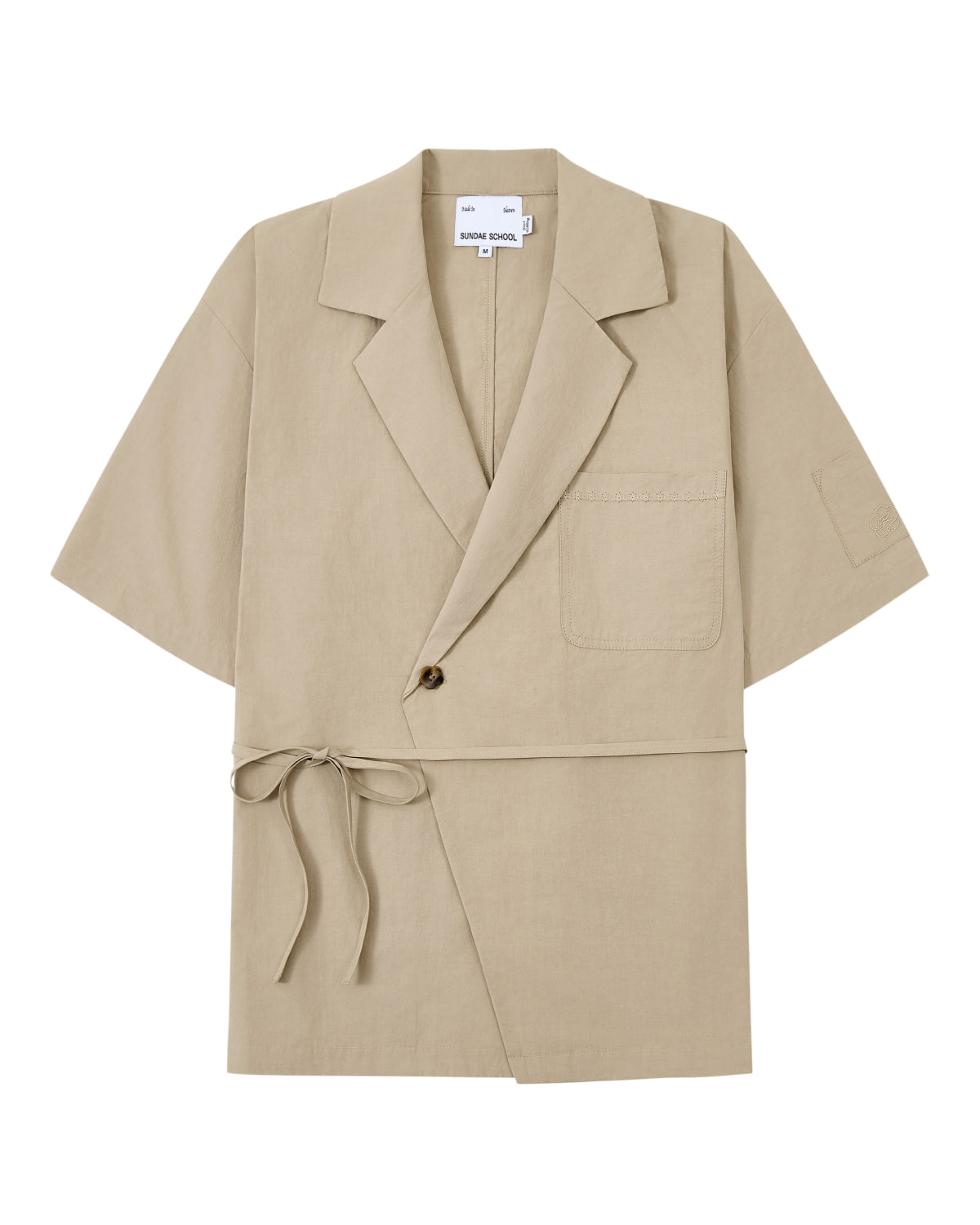 The WHEAT HANBOK LINEN SHIRT by Sundae School is a beige short-sleeve jacket made from a breathable cotton-linen blend, featuring a notched collar, single chest pocket, one-button closure, and side tie detail inspired by the modern hanbok shirt, shown laid flat on a white background.