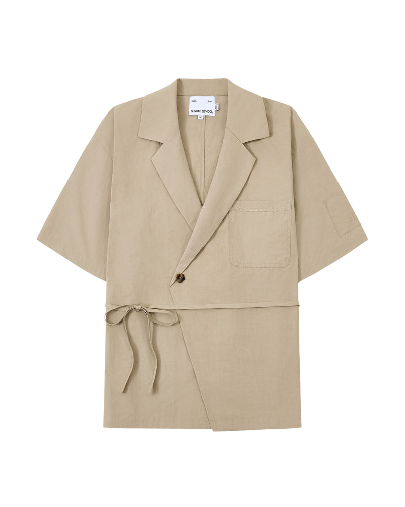 The WHEAT HANBOK LINEN SHIRT by Sundae School is a beige short-sleeve jacket made from a breathable cotton-linen blend, featuring a notched collar, single chest pocket, one-button closure, and side tie detail inspired by the modern hanbok shirt, shown laid flat on a white background.