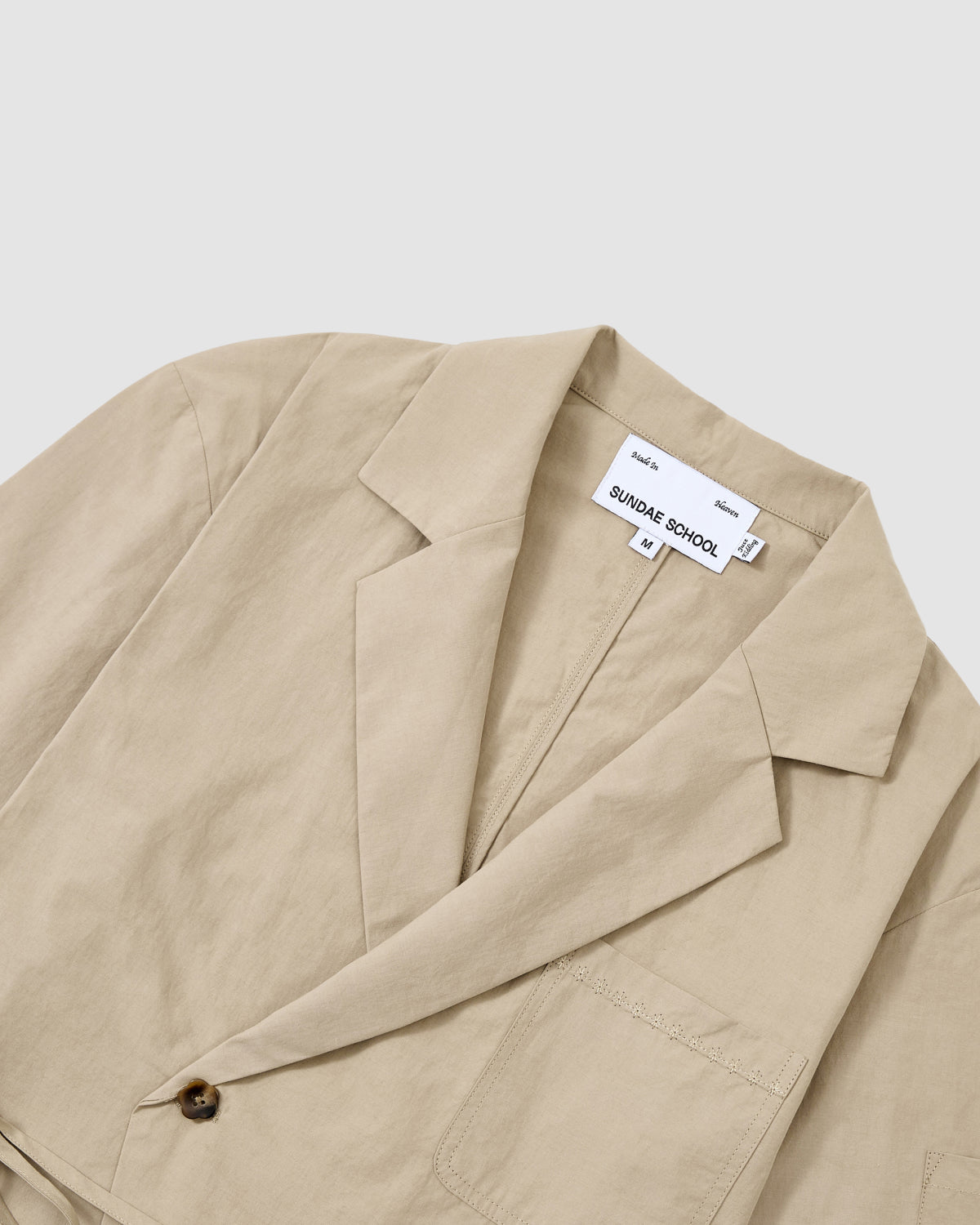 A close-up of the WHEAT HANBOK LINEN SHIRT by Sundae School, featuring a notched lapel, chest pocket, and single brown button in a cotton linen blend. Displayed flat with the label 