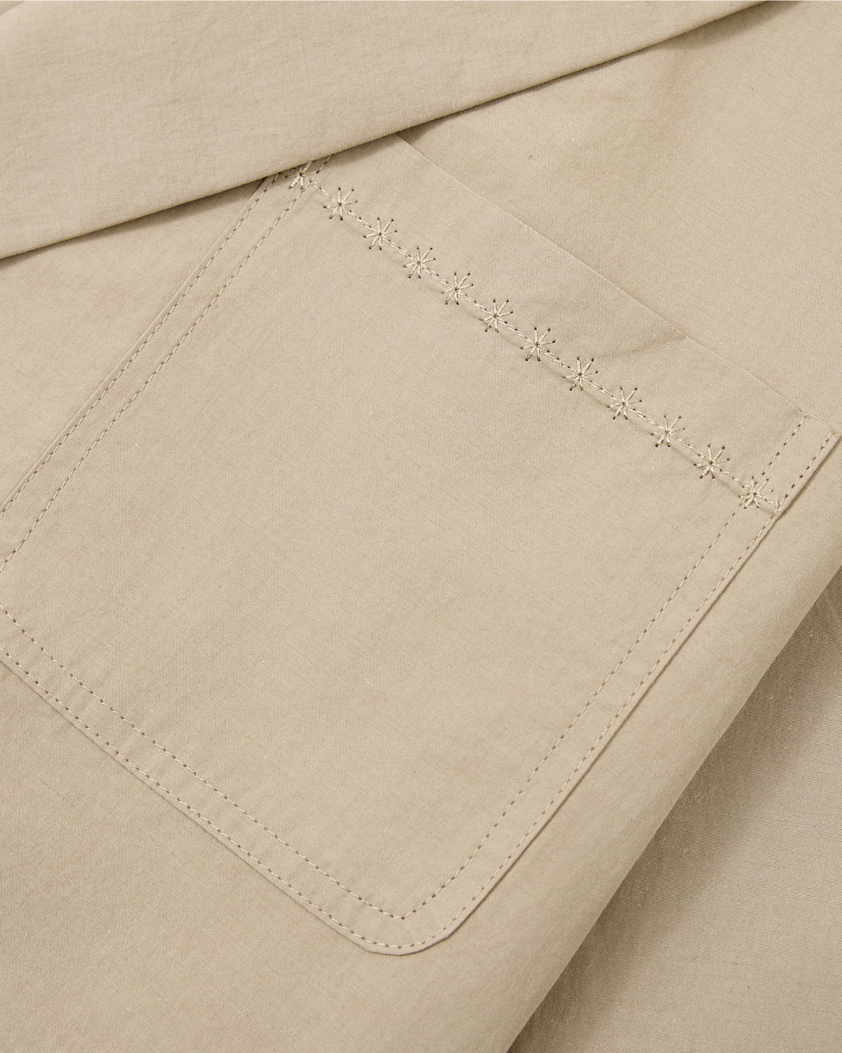 Close-up of the beige cotton linen blend fabric of the Sundae School WHEAT HANBOK LINEN SHIRT, featuring a square pocket. The pocket is detailed with a row of delicate, light-colored floral embroidery along the top edge, adding a subtle charm typical of summer shirts.