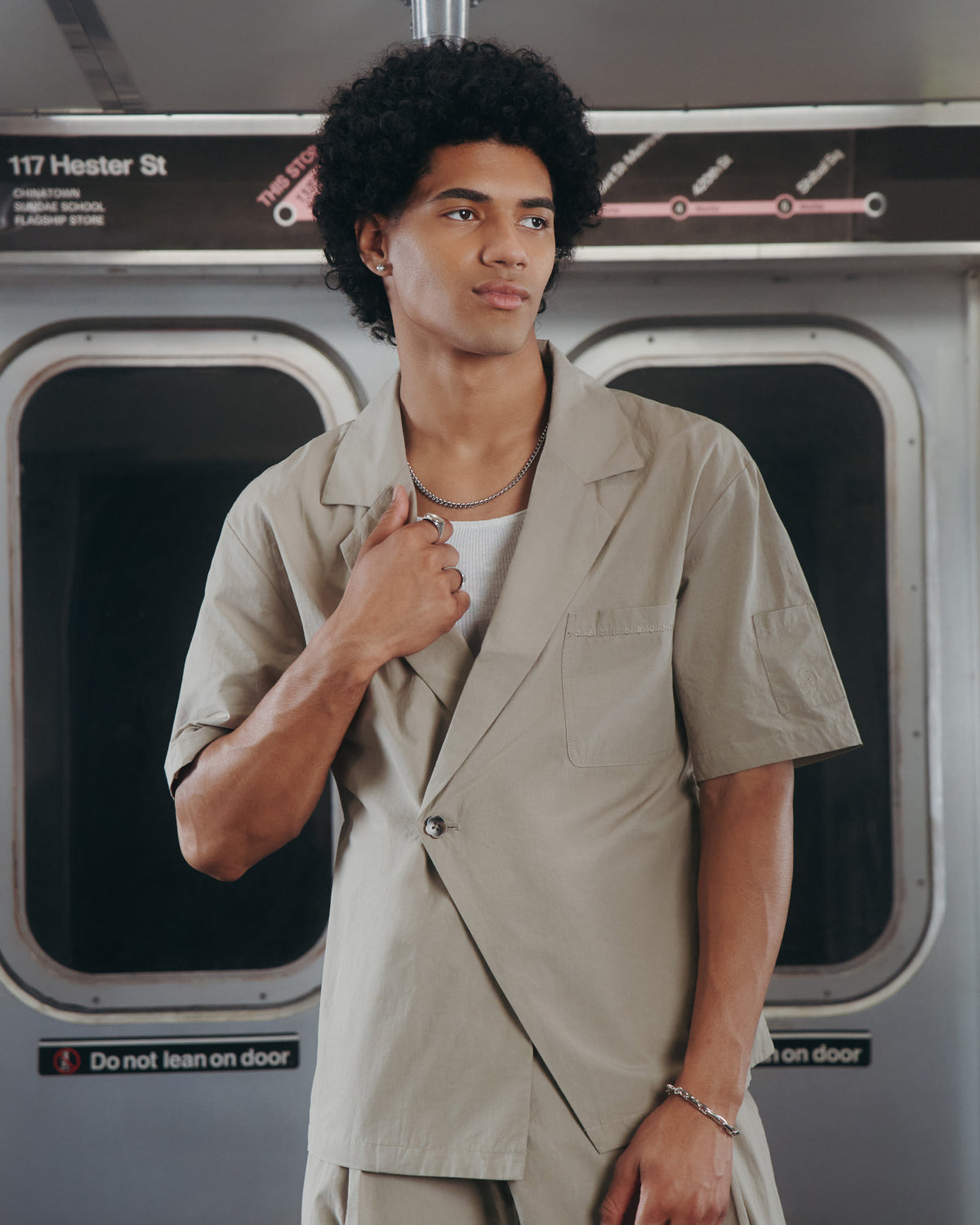 A person with curly hair, wearing the Sundae School WHEAT HANBOK LINEN SHIRT, stands in a subway car holding the lapel. A subway map and a sign reading "117 Hester St" are visible above the train doors.