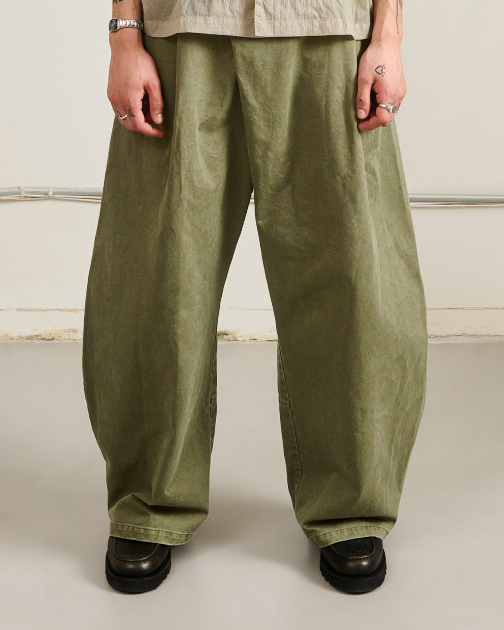 A person wearing very loose, wide-leg olive green pants with black shoes, shown from the waist down against a plain light beige studio background.