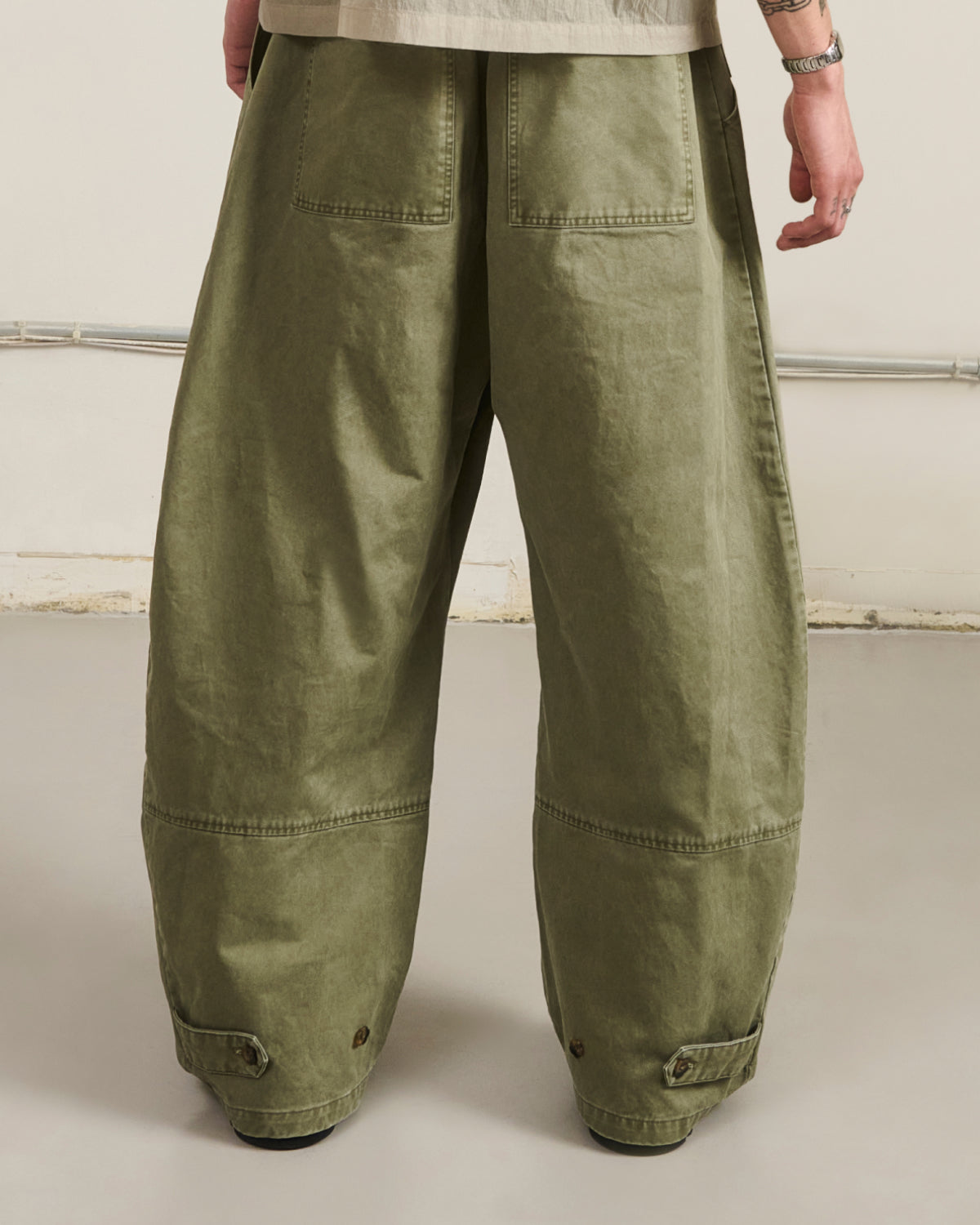 A person wearing very baggy olive green cargo pants, shown from the back against a plain off-white studio wall.
