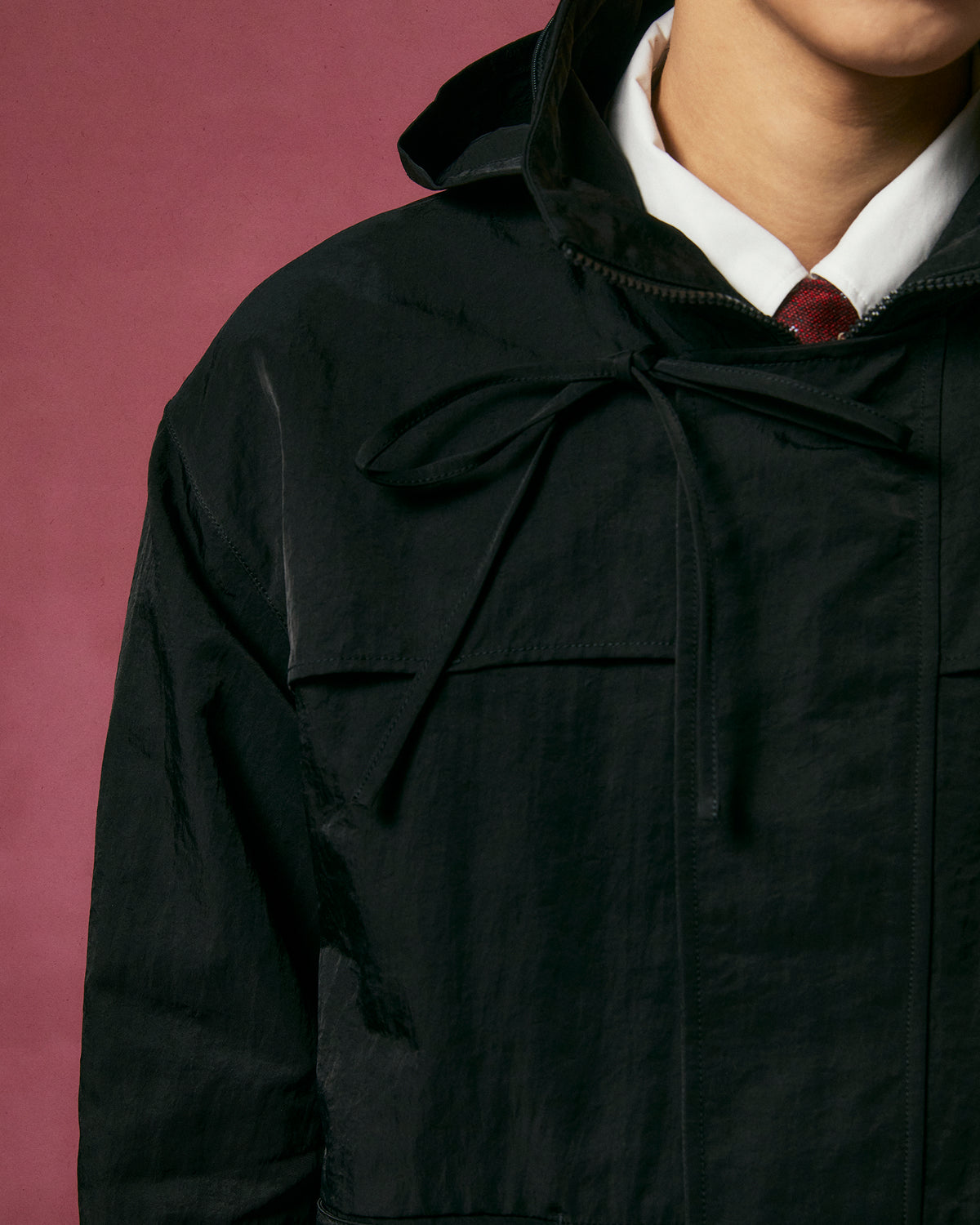 A person wears the Sundae School BLACK HANBOK TRACK JACKET over a white collared shirt and red tie, set against a dark pink background. The cropped image shows only the lower face and upper torso.