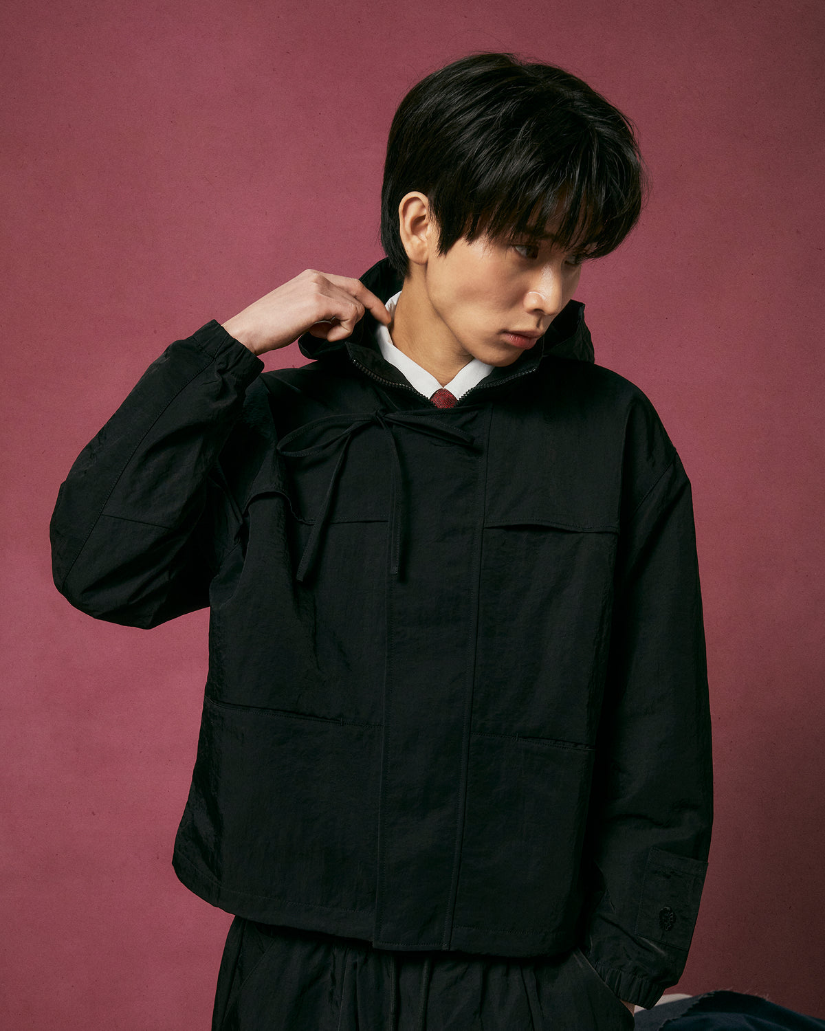 A person with short dark hair, looking to the side and touching the collar, wears the BLACK HANBOK TRACK JACKET by Sundae School against a plain dark pink background.