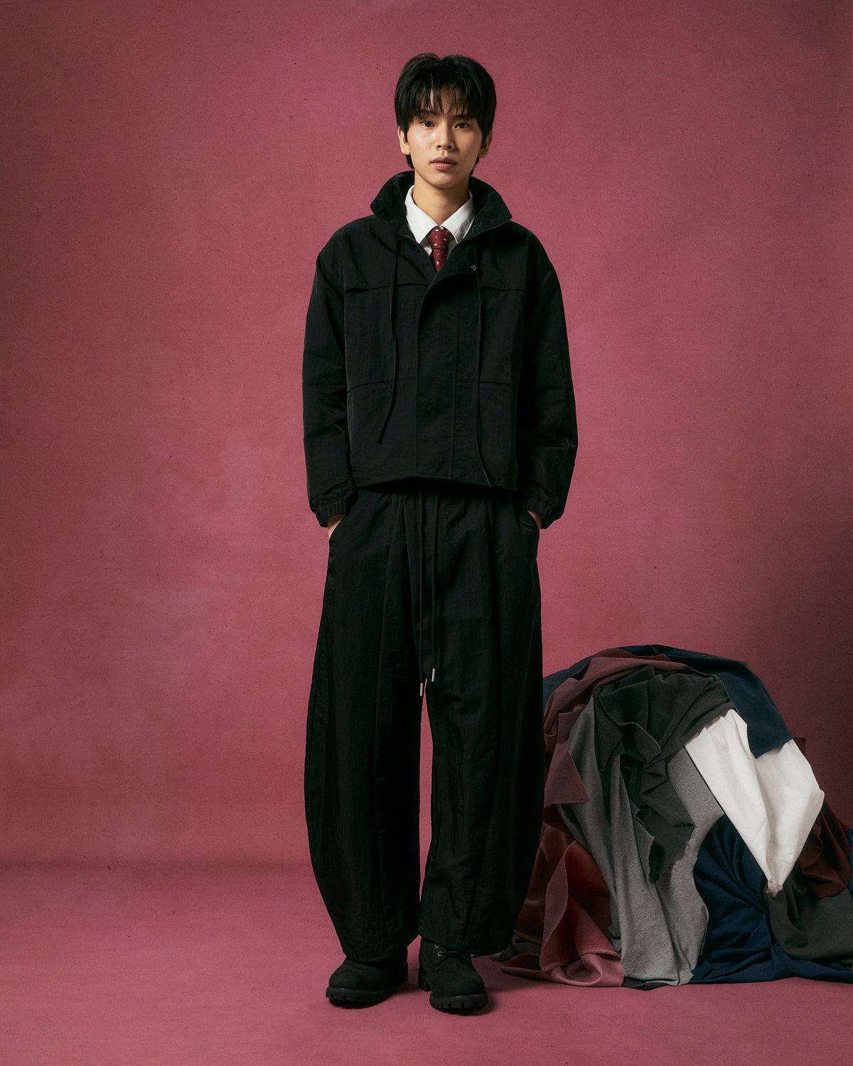 A person stands against a mauve backdrop wearing the BLACK HANBOK TRACK JACKET by Sundae School, paired with loose black pants, black shoes, a white shirt, and a red tie. Colorful folded and draped fabrics are arranged to their right.