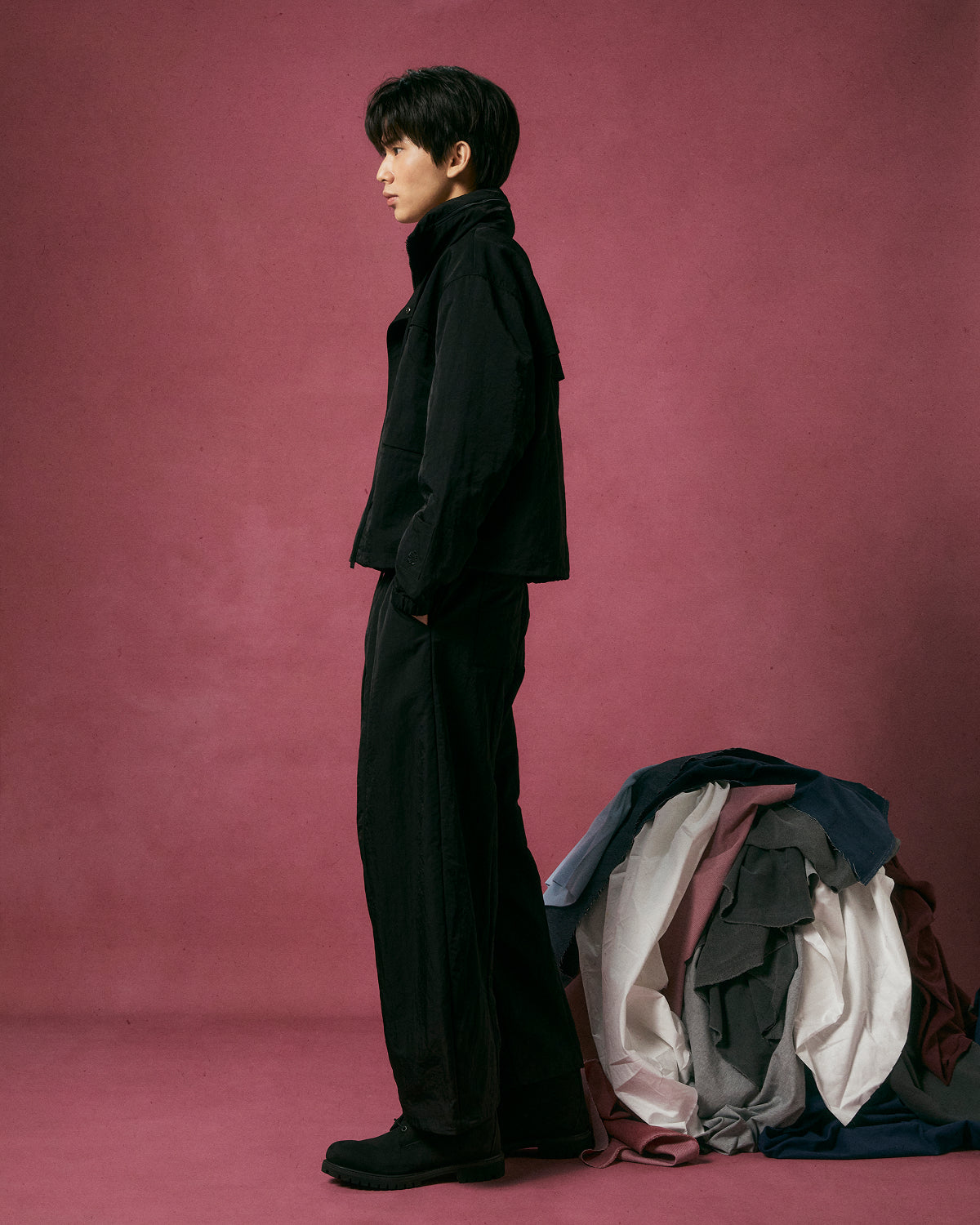 A person wearing Sundae School’s BLACK HANBOK BALLOON TRACK PANTS stands in profile against a pink backdrop, hands in pockets, beside a large pile of colorful fabrics on the floor.