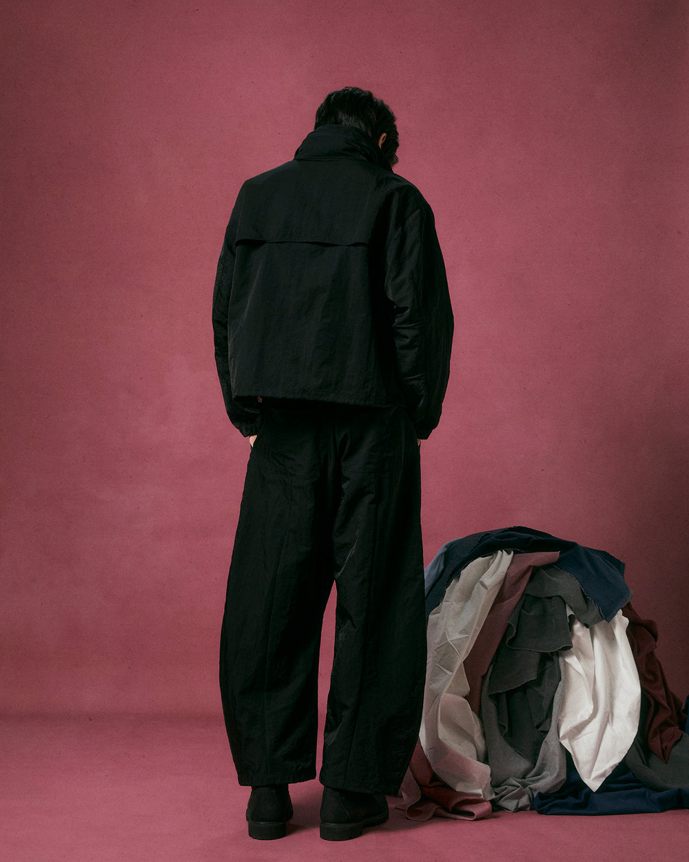 Wearing the Sundae School BLACK HANBOK TRACK JACKET, a person in loose black clothing stands with their back to the camera against a pink background, next to a pile of assorted colored fabrics on the floor.