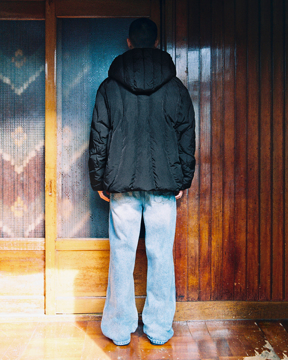 A person wearing the Sundae School BLACK HEAVYWEIGHT MOCHI PUFFER, a black hooded jacket filled with duck down, and light blue jeans stands indoors facing a wooden wall with sliding glass doors, their back to the camera.