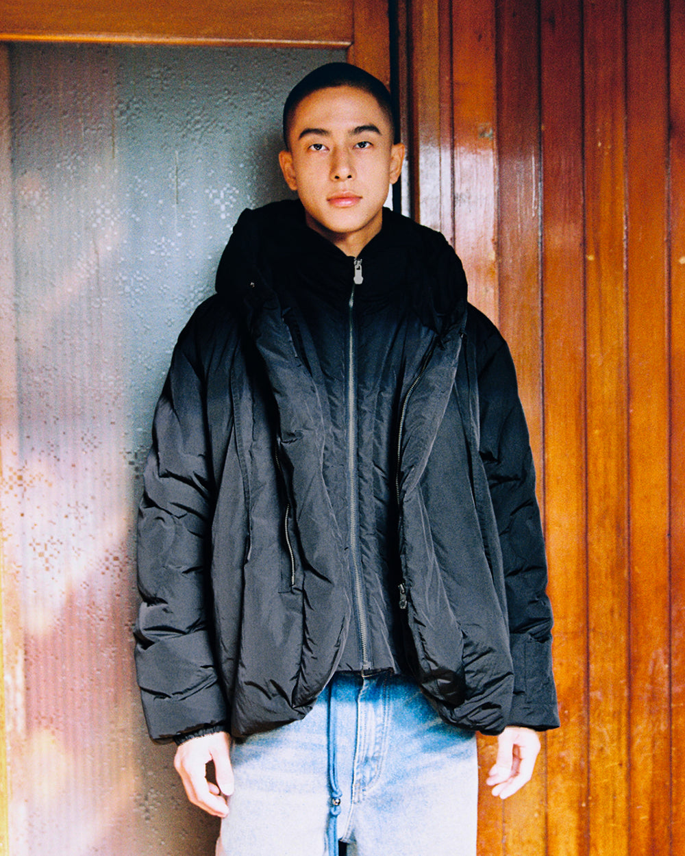 A person with short hair, dressed in the Sundae School BLACK HEAVYWEIGHT MOCHI PUFFER and blue jeans, stands in front of a wooden door and a frosted glass panel, looking directly at the camera with a neutral expression.