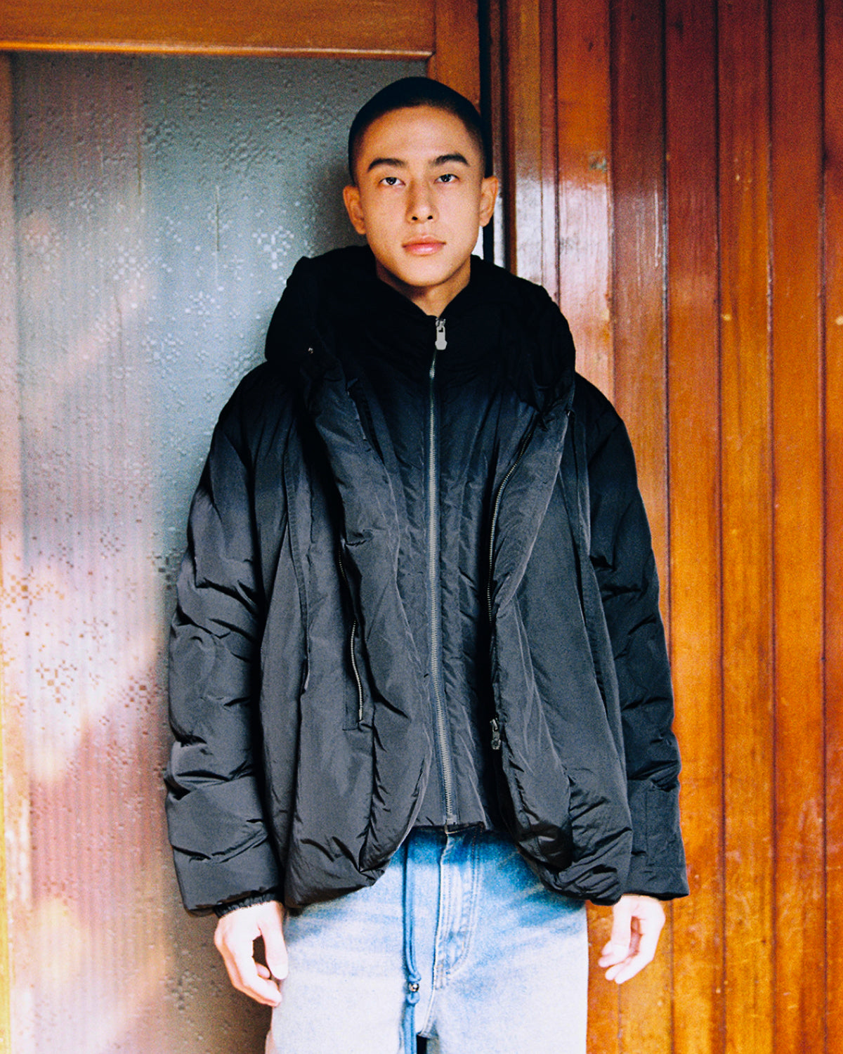 A person with short hair, dressed in the Sundae School BLACK HEAVYWEIGHT MOCHI PUFFER and blue jeans, stands in front of a wooden door and a frosted glass panel, looking directly at the camera with a neutral expression.
