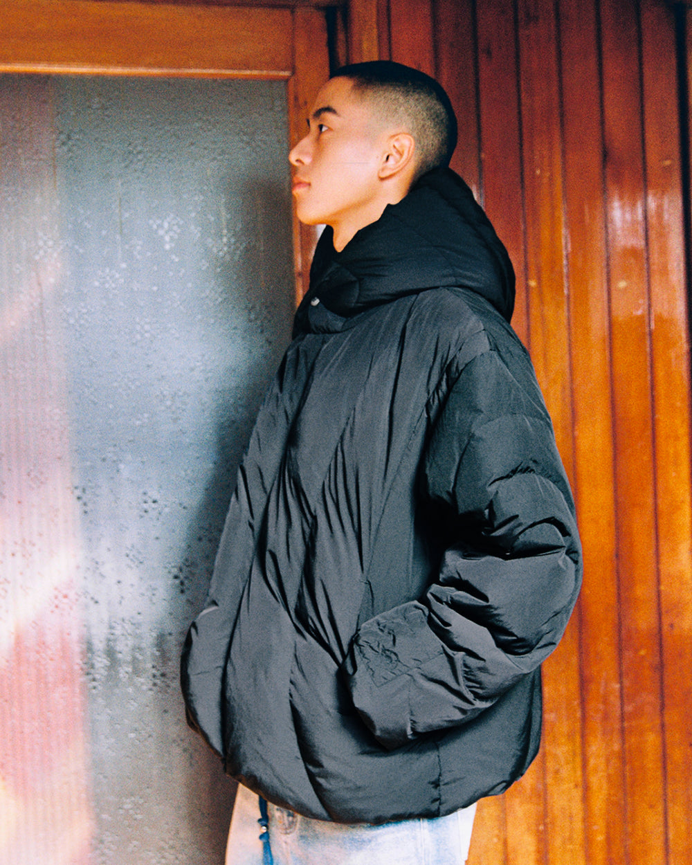 Wearing the Sundae School BLACK HEAVYWEIGHT MOCHI PUFFER, a person stands in profile against a wooden wall and frosted glass door, hands in pockets and looking upward.