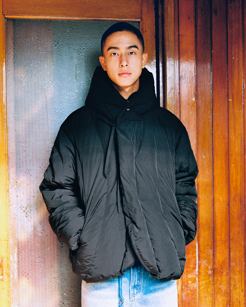 A person wearing the Sundae School BLACK HEAVYWEIGHT MOCHI PUFFER with a hood and light blue jeans stands in front of a wooden door, hands in pockets, looking directly at the camera.