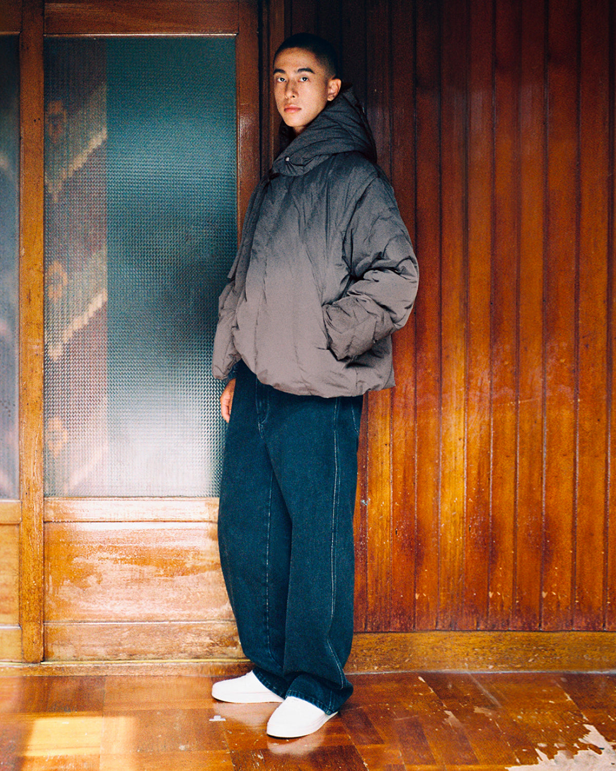 Indoors, a person is seen wearing Sundae School’s CHARCOAL HEAVYWEIGHT MOCHI PUFFER—a cozy duck down-filled jacket—styled with wide dark blue jeans and white shoes. The warm, rustic ambiance of the wooden wall and door forms a welcoming backdrop.
