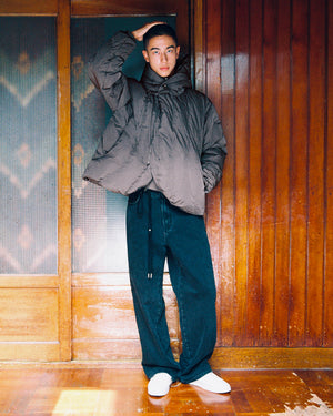 A person in the Sundae School CHARCOAL HEAVYWEIGHT MOCHI PUFFER, loose dark jeans, and white shoes stands indoors against wooden paneling, one hand touching their head and the other in their pocket.