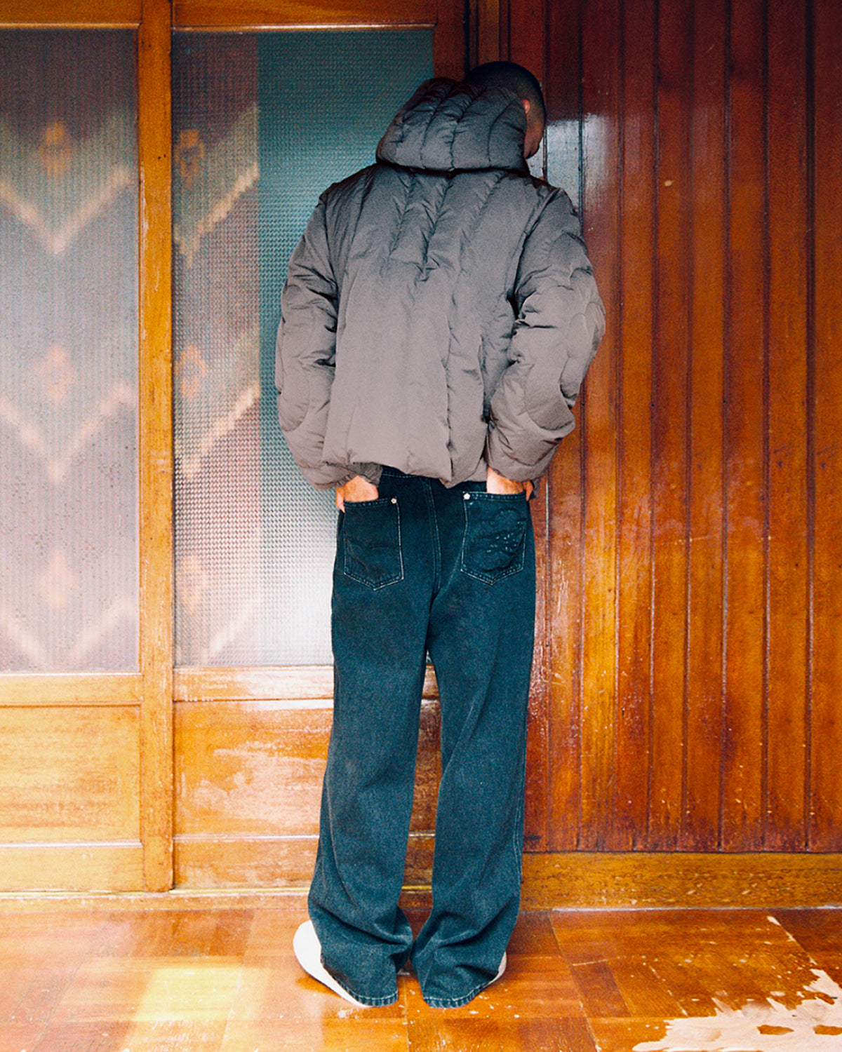 A person wearing the CHARCOAL HEAVYWEIGHT MOCHI PUFFER by Sundae School and dark jeans stands with their back to the camera facing a wooden wall, head turned slightly to the side.