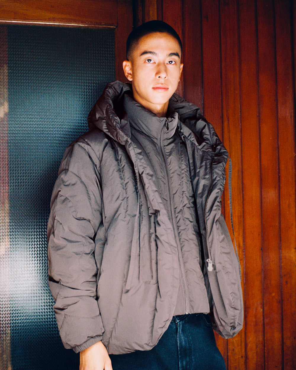 A person with short hair stands indoors, facing the camera with a neutral expression, wearing Sundae School’s CHARCOAL HEAVYWEIGHT MOCHI PUFFER and dark pants. The background features wooden paneling and a frosted glass door.