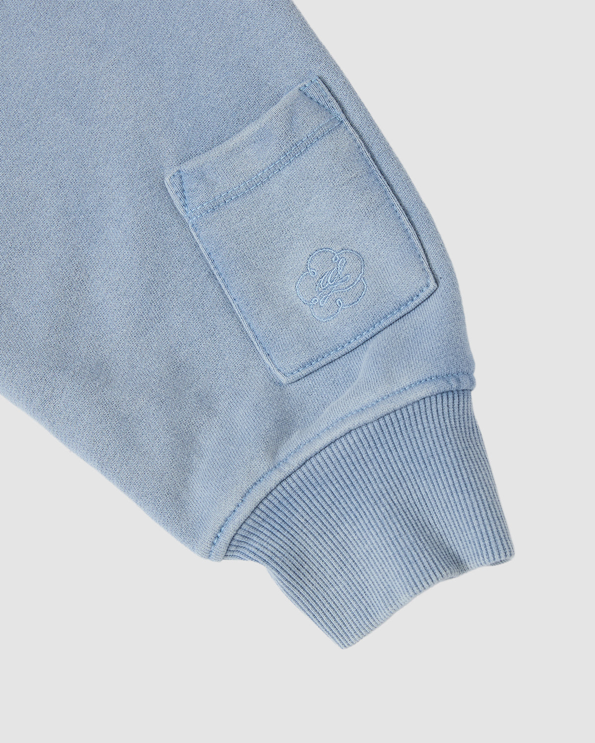 Close-up of the SKY BLUE HESTER QUARTER ZIP by Sundae School, showing its light blue cotton sleeve with a small square stash pocket and embroidered floral logo above the ribbed cuff.