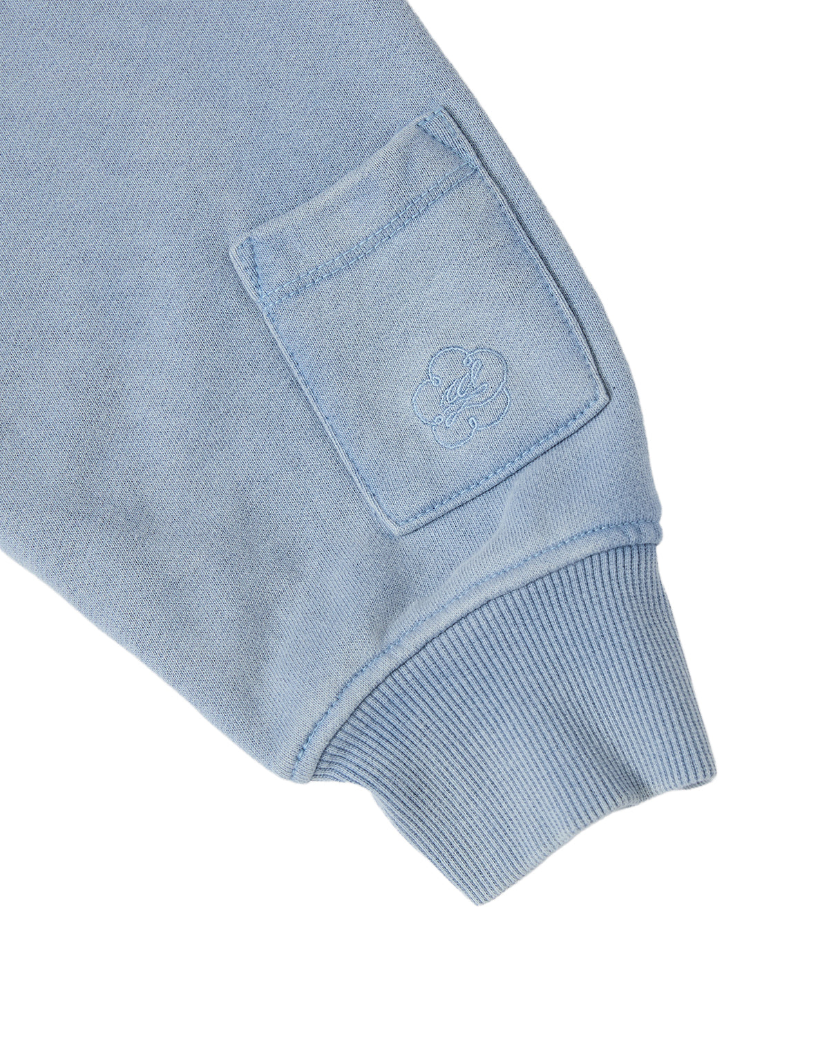 The SKY BLUE HESTER QUARTER ZIP from Sundae School features a soft, slightly textured cotton fabric in a light blue hue, with pullover sleeves finished with ribbed cuffs and a small square pocket adorned with an embroidered flower design.