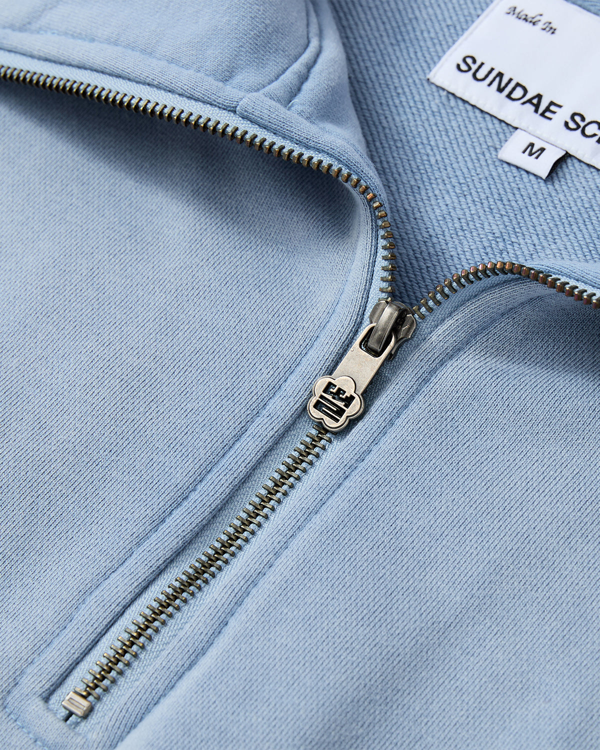 Close-up of the SKY BLUE HESTER QUARTER ZIP by Sundae School, featuring an oversized fit in a light blue shade. The quarter zip is partially unzipped to reveal a metallic zipper and a white label with Sundae School branding and size M. The soft, textured fabric is paired with a relaxed funnel neck collar for extra comfort.
