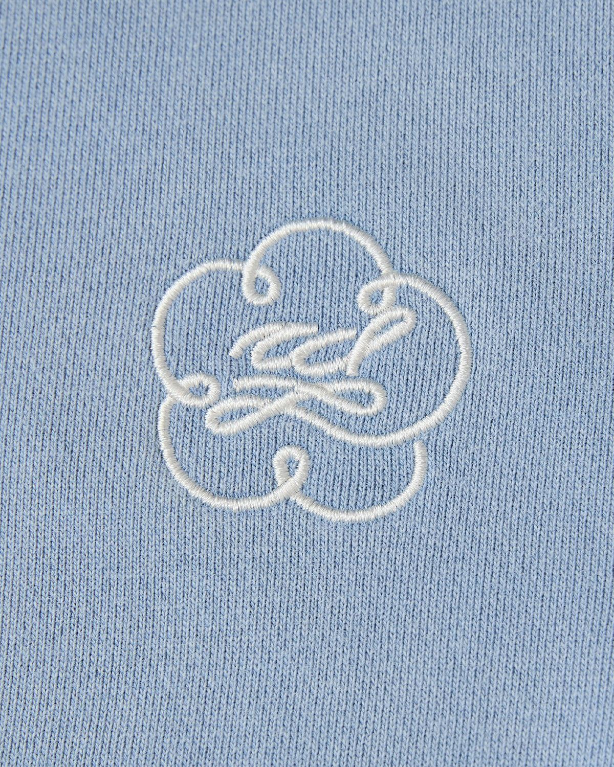 A close-up of the blue fabric from Sundae School’s SKY BLUE HESTER QUARTER ZIP, showcasing a white abstract embroidered design reminiscent of a stylized cloud or flower.