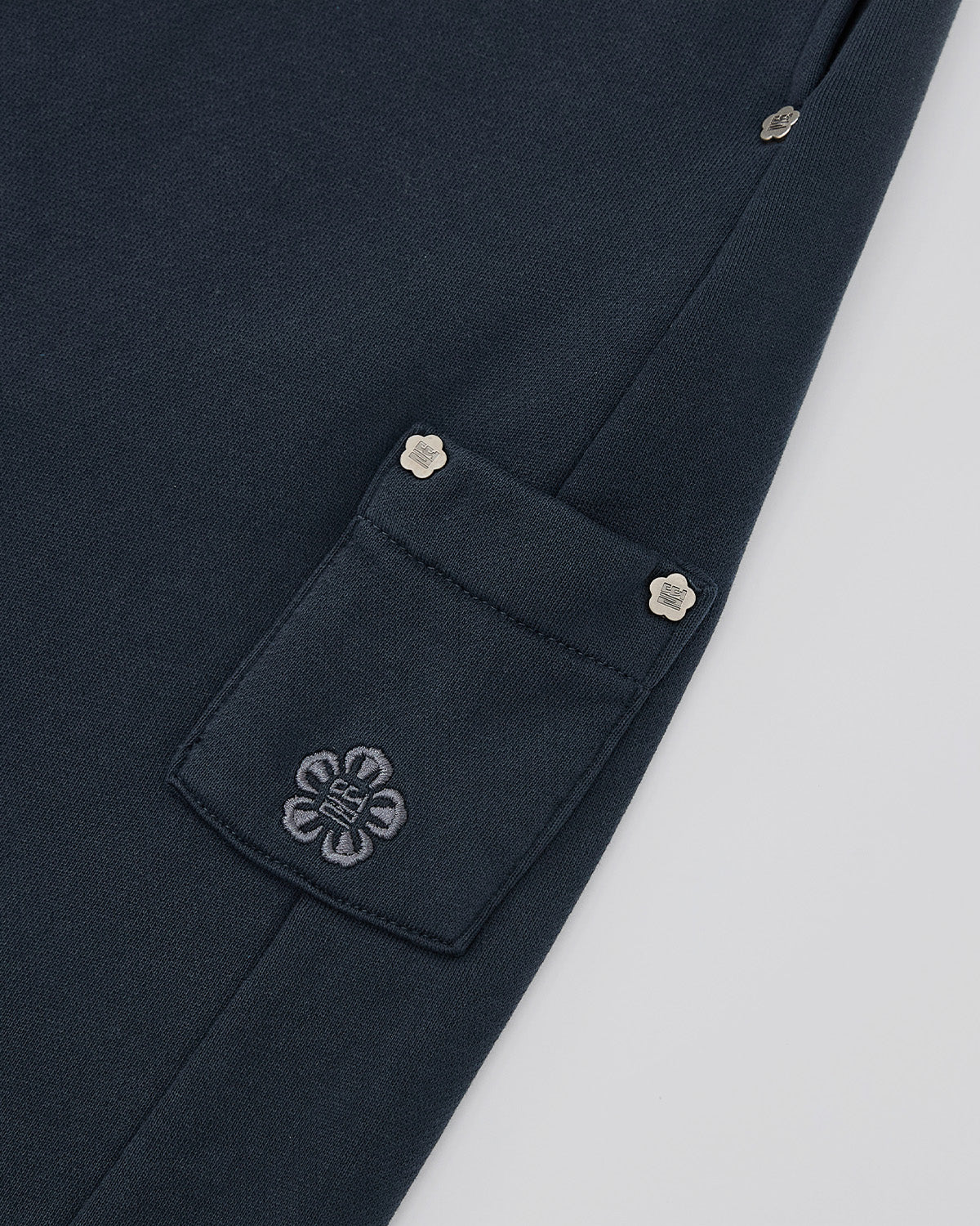 Close-up of Sundae School’s INDIGO WASH WRAP SWEATBAJI: dark navy fabric with a pocket, silver flower-shaped button, and an embroidered floral logo. The seam and matching buttons echo the elegant lines of hanbok-inspired wide leg pants.