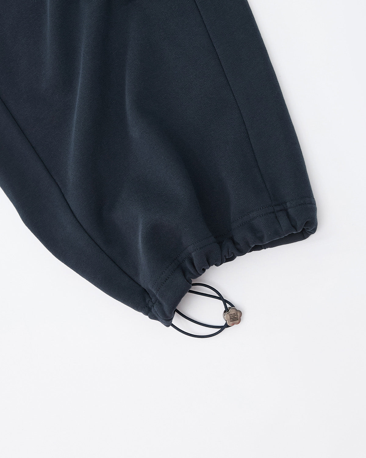 A close-up of the INDIGO WASH WRAP SWEATBAJI by Sundae School shows its dark, hanbok-inspired pant leg with an elastic drawstring and cord lock at the hem, set against a white background.