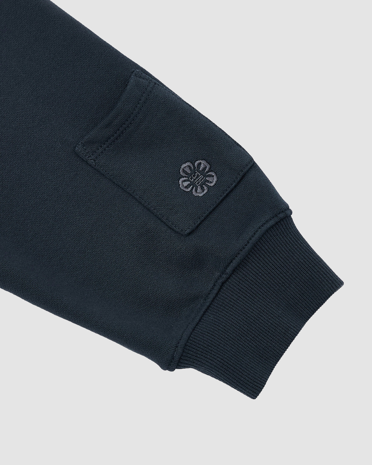 Close-up of Sundae School's INDIGO WASH HANBOK HOODIE sleeve in dark navy, showing a ribbed cuff, small rectangular patch, embroidered clover-like detail near the patch, and a hidden signature sleeve stash pocket. Plain light gray background.