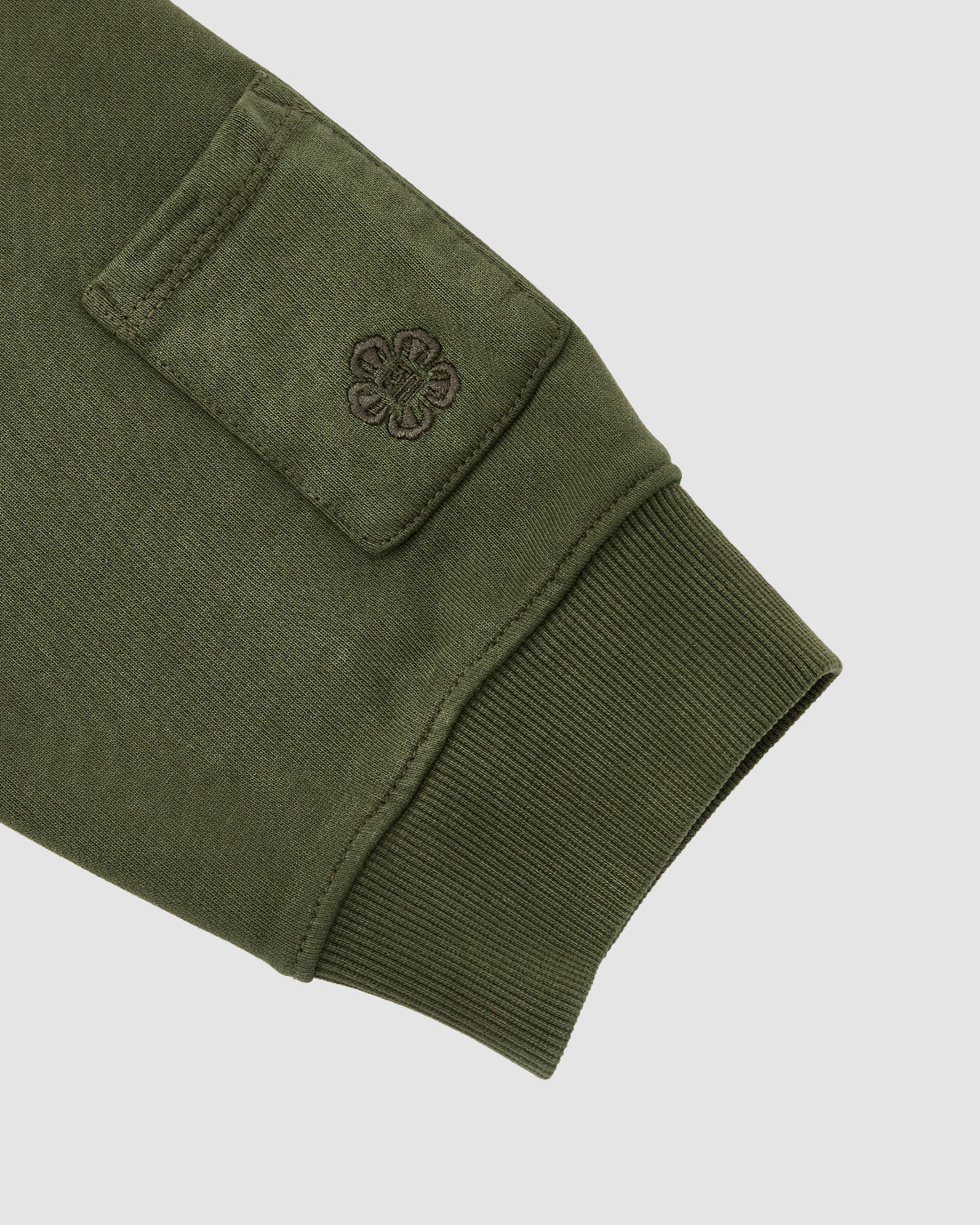 Close-up of the KHAKI HANBOK HOODIE by Sundae School, showing an olive green sleeve with a ribbed cuff and a stash pocket embroidered with a flower. The background is light gray.