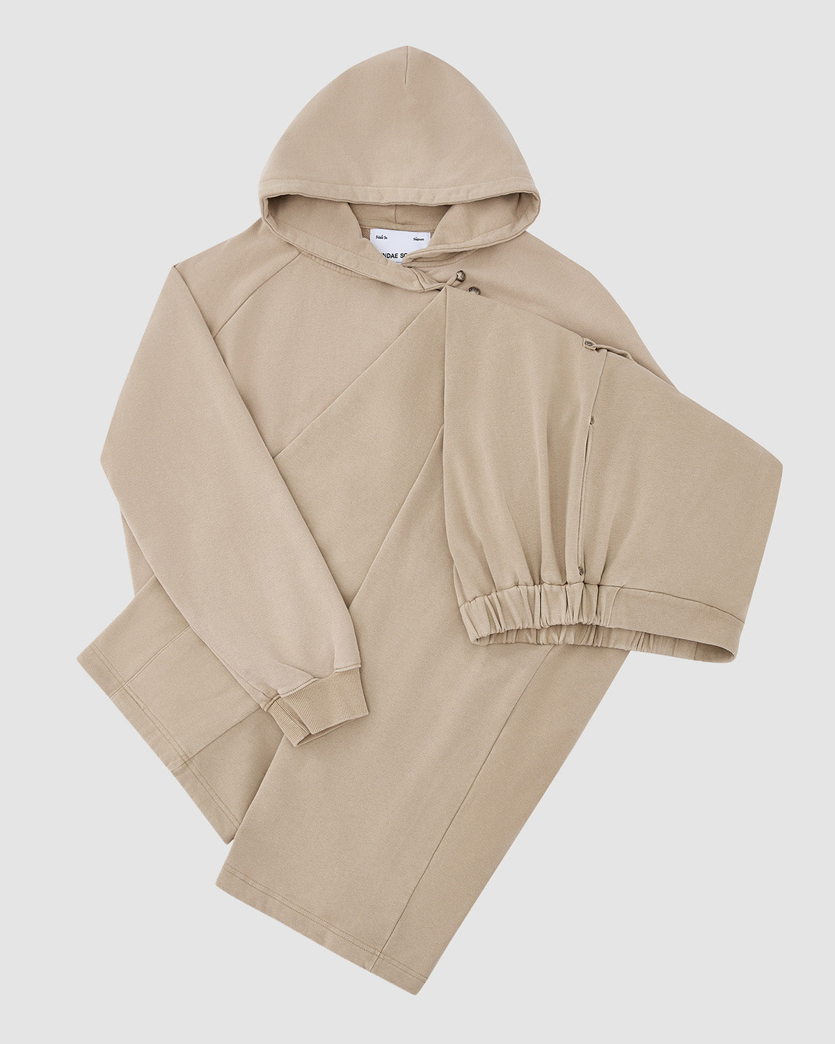 The TAUPE WRAP SWEATBAJI by Sundae School, a beige 100% cotton hoodie with long sleeves, angled seams, and elastic cuffs, is shown flat on a white background with one sleeve folded across the front.