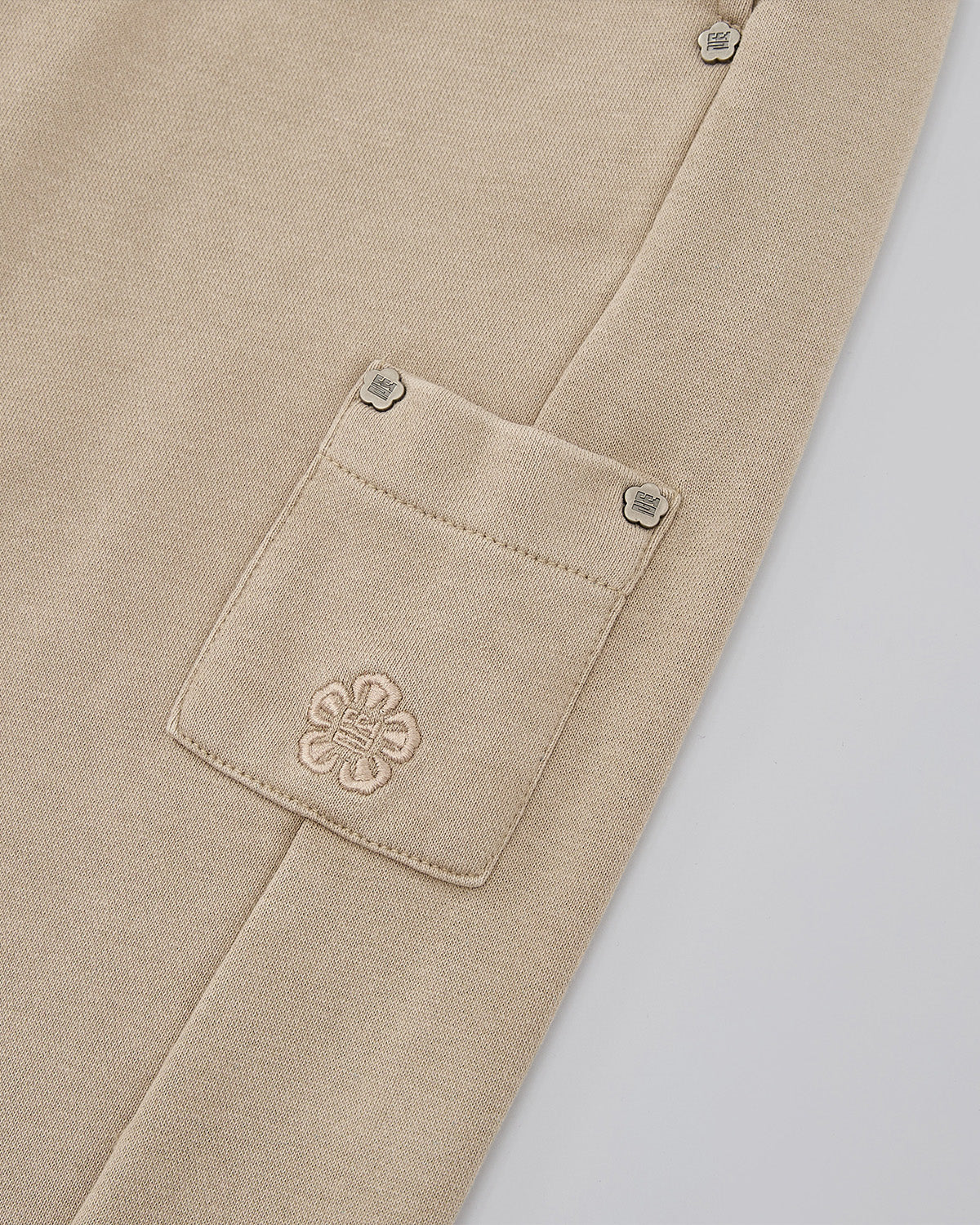 Close-up of the TAUPE WRAP SWEATBAJI by Sundae School: a beige 100% cotton garment with textured fabric, featuring a small rectangular pocket with embroidered floral design and two metallic snap buttons on the flap.