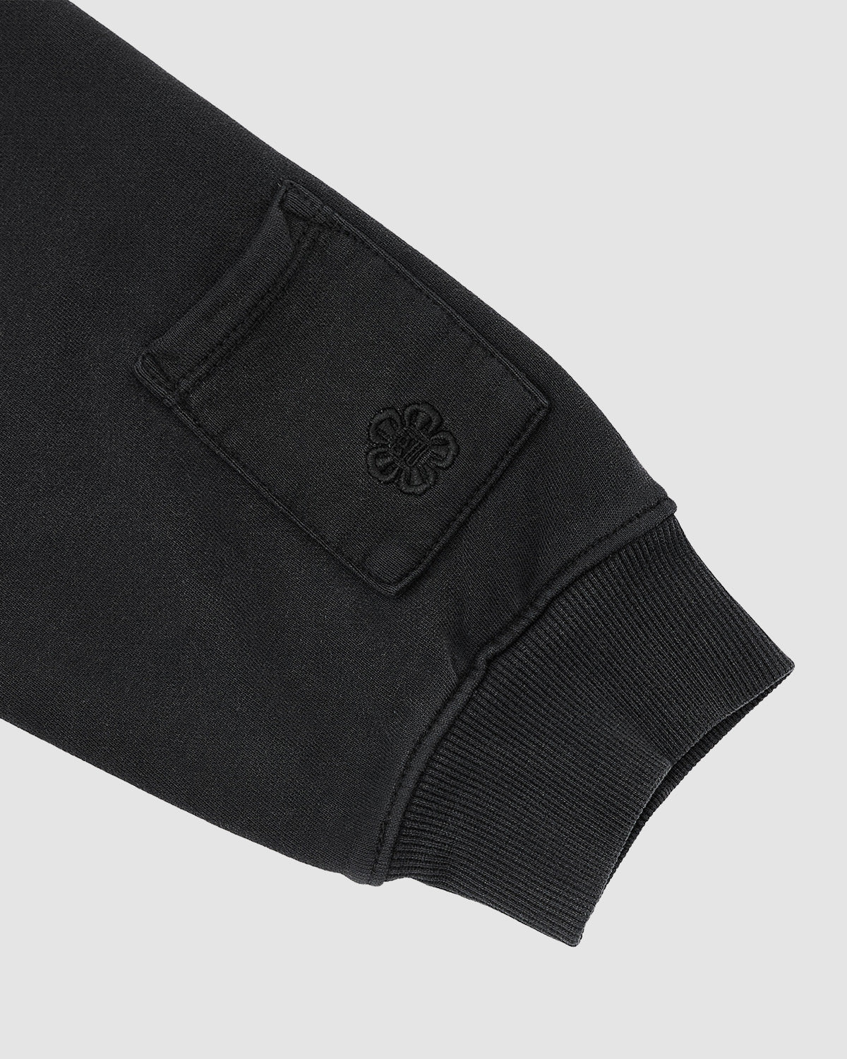 The FADE BLACK HANBOK HOODIE by Sundae School features black long sleeves with ribbed cuffs and a small rectangular stash pocket above the wrist, embroidered with a flower-like design, set against a light gray background.