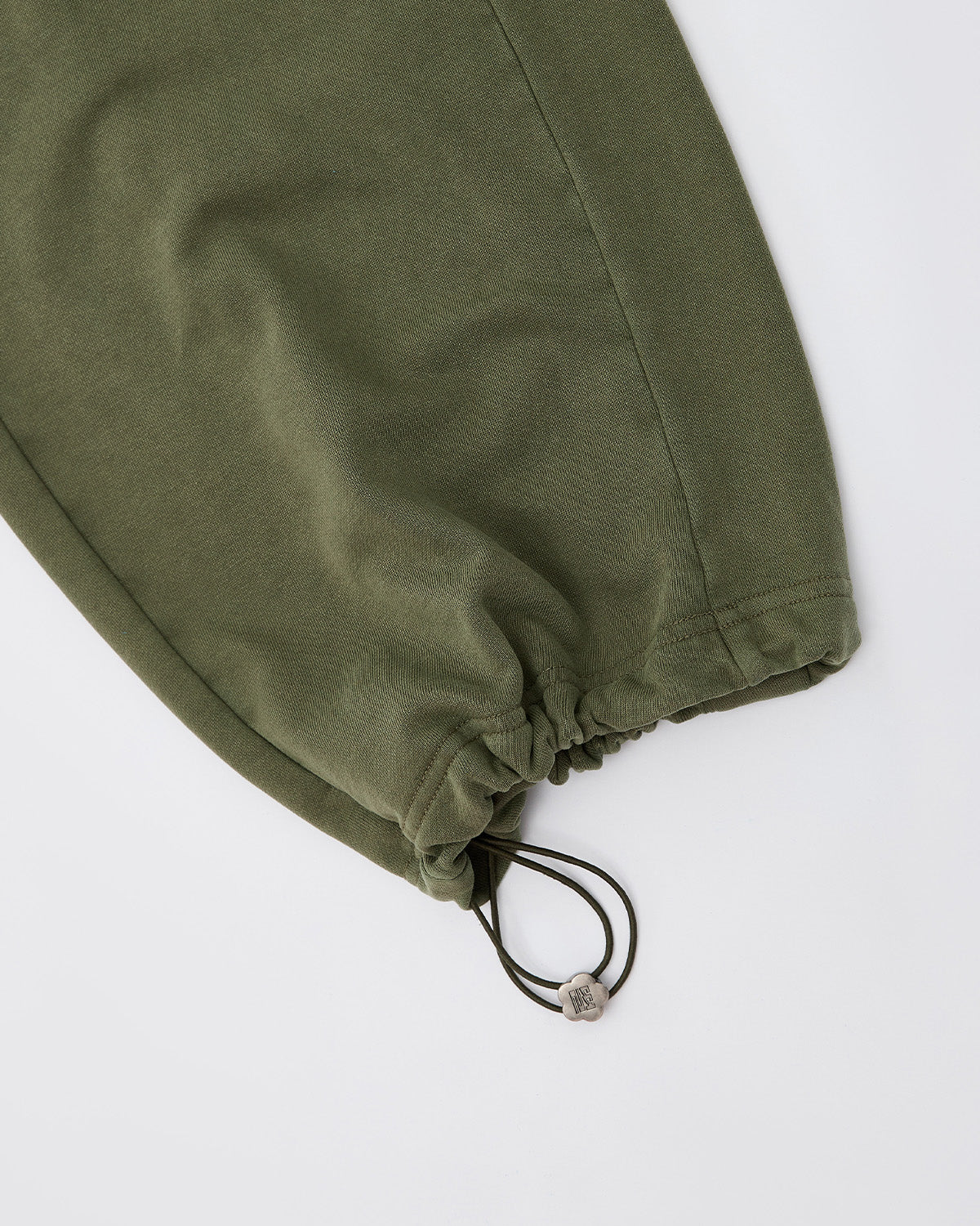 Close-up of the Sundae School KHAKI WRAP SWEATBAJI pant leg in olive green, highlighting the Hanbok-inspired adjustable drawstring hem with two cords and a small bead, laid flat on a white surface.