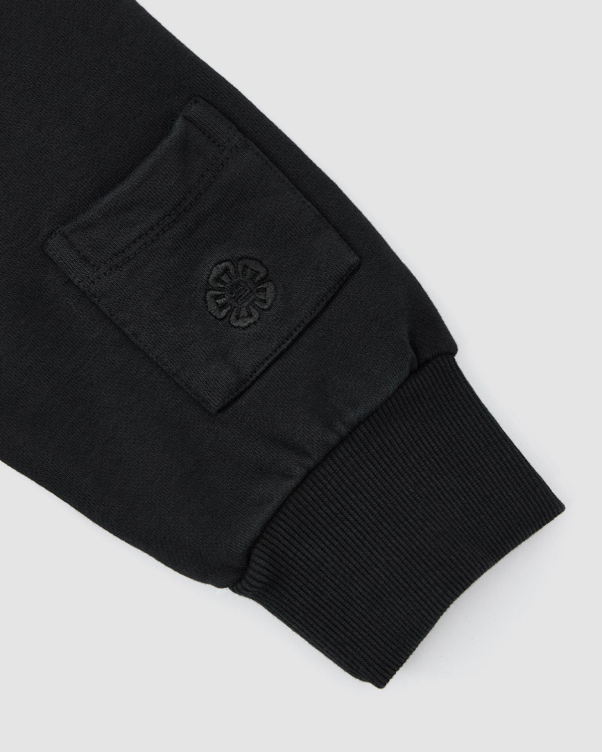 A close-up of the sleeve on the Sundae School HIGH HORSE LNY HOODIE shows a ribbed cuff, a small pocket, and an embroidered flower-like emblem on the pocket.