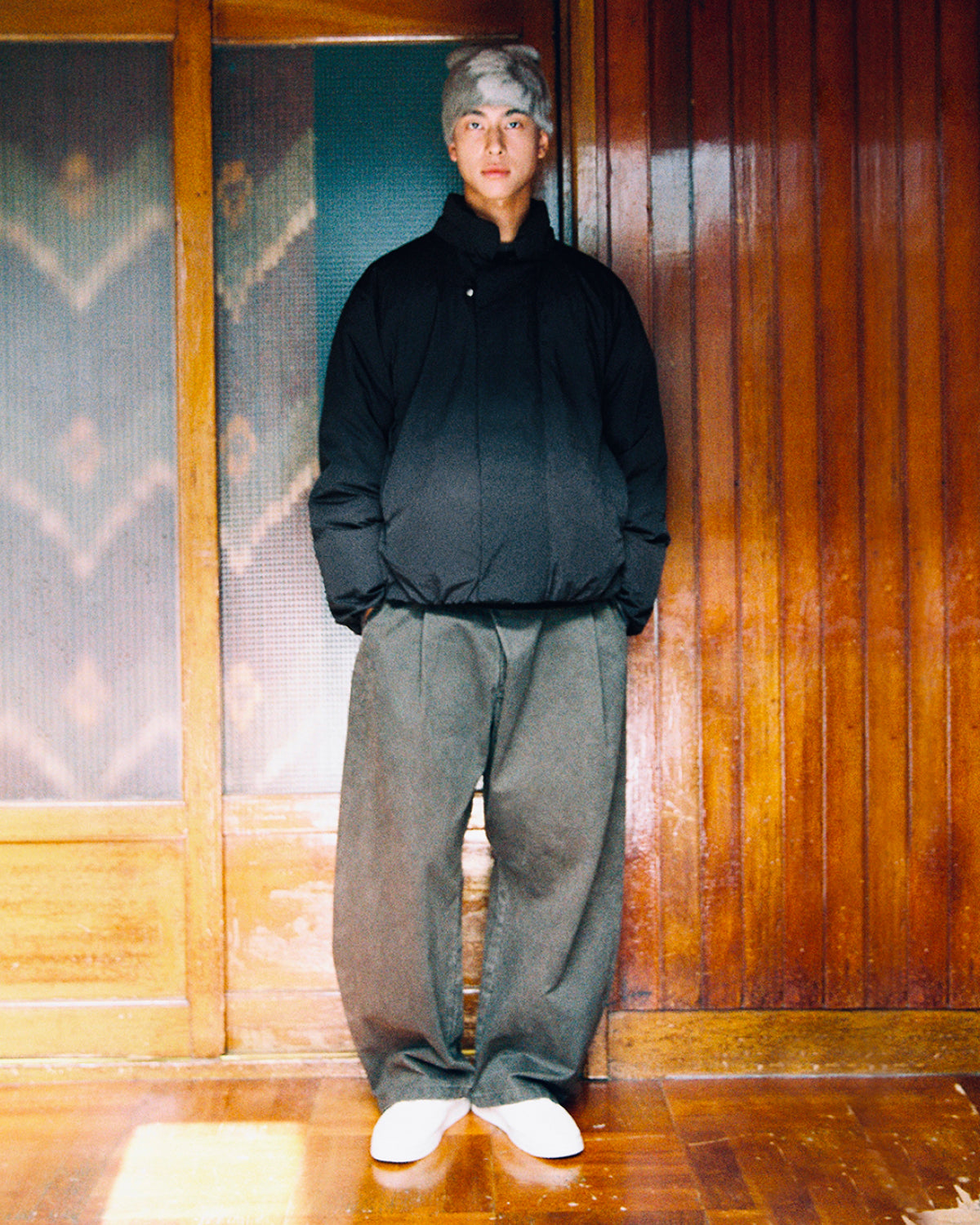 A person with light hair stands indoors against wood paneling, wearing Sundae School’s BLACK IBUL DOWN PUFFER, paired with wide gray pants and white shoes, looking straight at the camera.