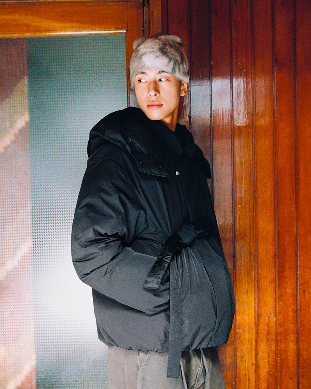 Wearing the BLACK IBUL DOWN PUFFER by Sundae School—an oversized black cocoon puffer coat with a detachable hood—a person stands indoors by a wooden wall, looking to the side and accessorized with a light gray furry hat.